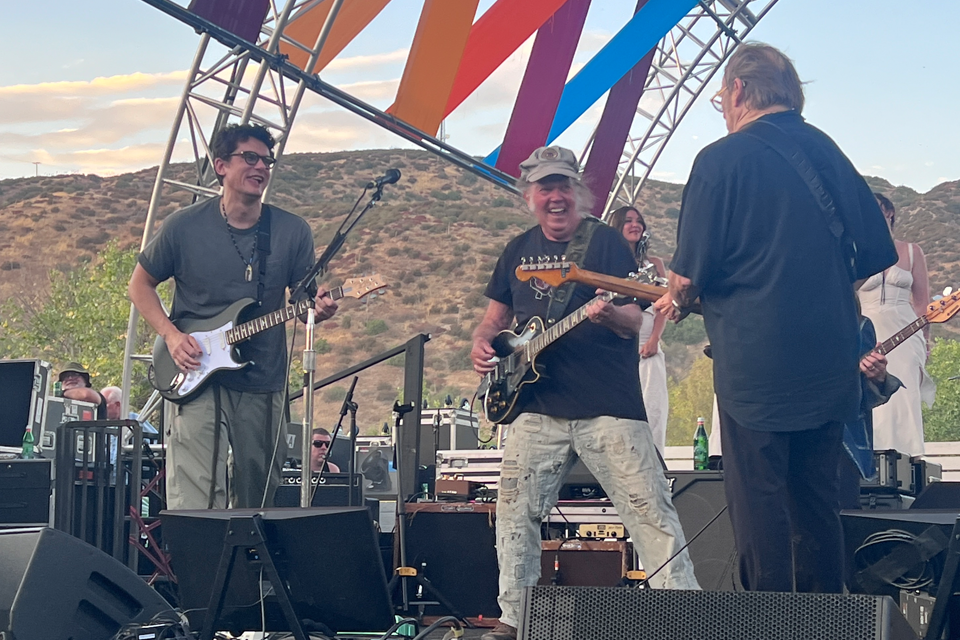 John Mayer, Neil Young, and Stephen Stills all playing guitars and smiling together on stage 