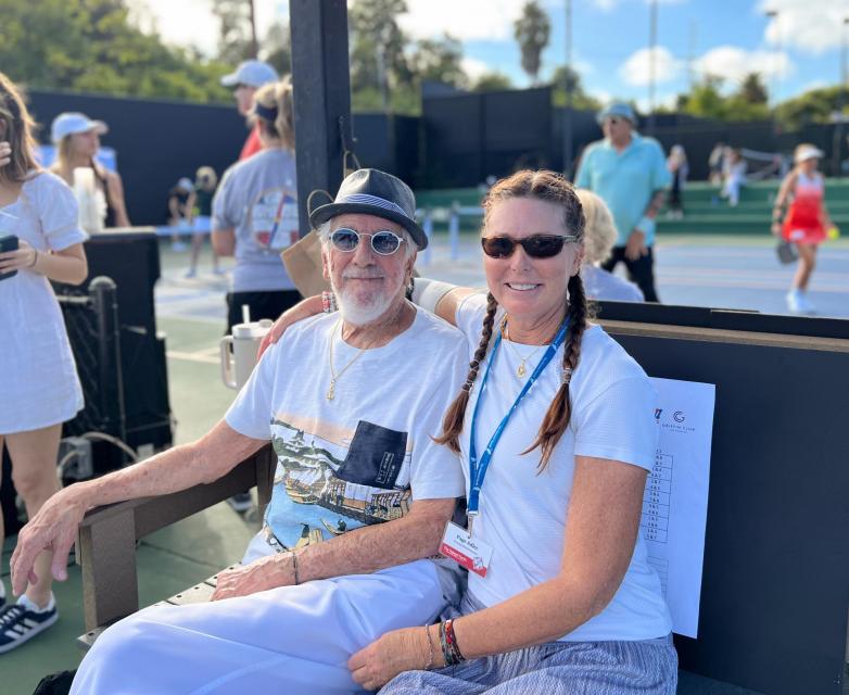 Lou and Page Adler sitting on a bench, courtside, watching the pickleball matches.