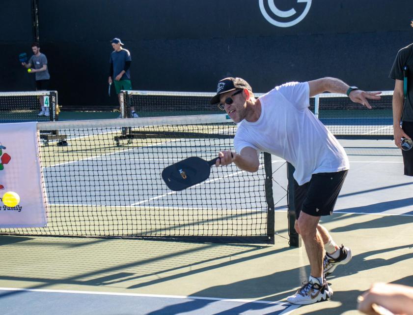Pickleball player swings his paddle at the ball mid air. 